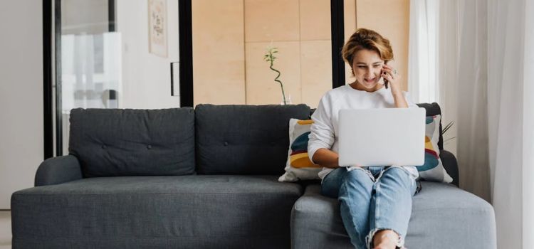 Mulher sentada no sofá com notebook no colo e telefone na mão